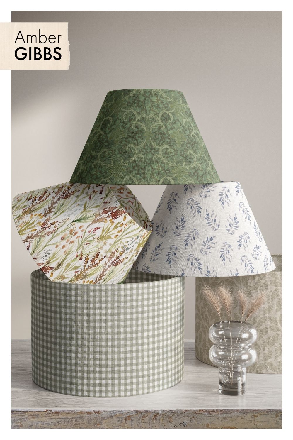 Stacked lampshade mockups showing five coordinating patterns: a sage green damask cone shade, a botanical wildflower print in rust and gold tones, a blue leaf sprig print on cream, a sage and white gingham drum shade, and a soft green floral drum shade in the background. Branded 'Amber Gibbs' logo in the top left corner, with a clear bubble vase holding dried pampas grass in front